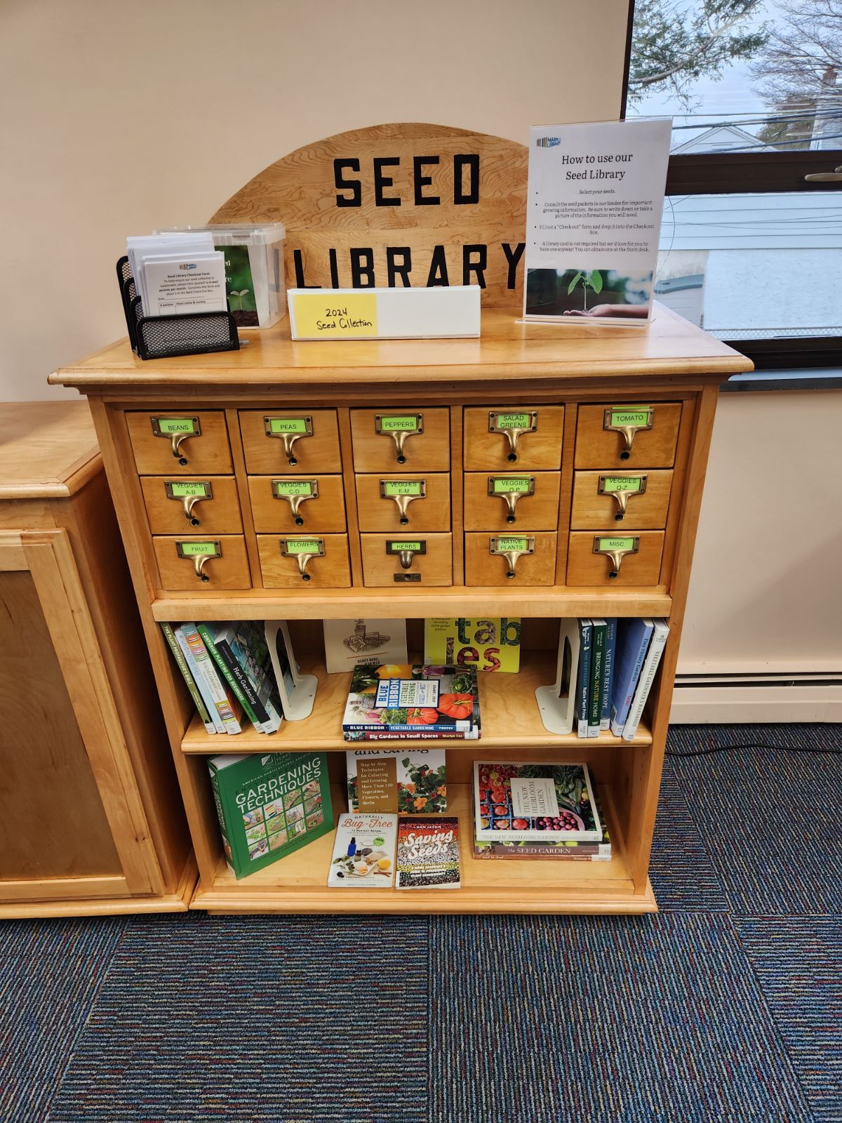 The Seed Library at Marple Library A wooden seed library cabinet with labeled drawers, plant and gardening books, brochures, and a sign with instructions on how to use the seed library.
